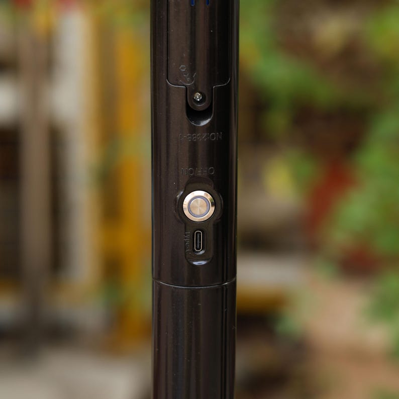 May include: Close-up of a black, cylindrical device with a silver button and a small rectangular port. The device has the word "OFF/ON" printed on it. The background is blurred, with green and yellow hues.