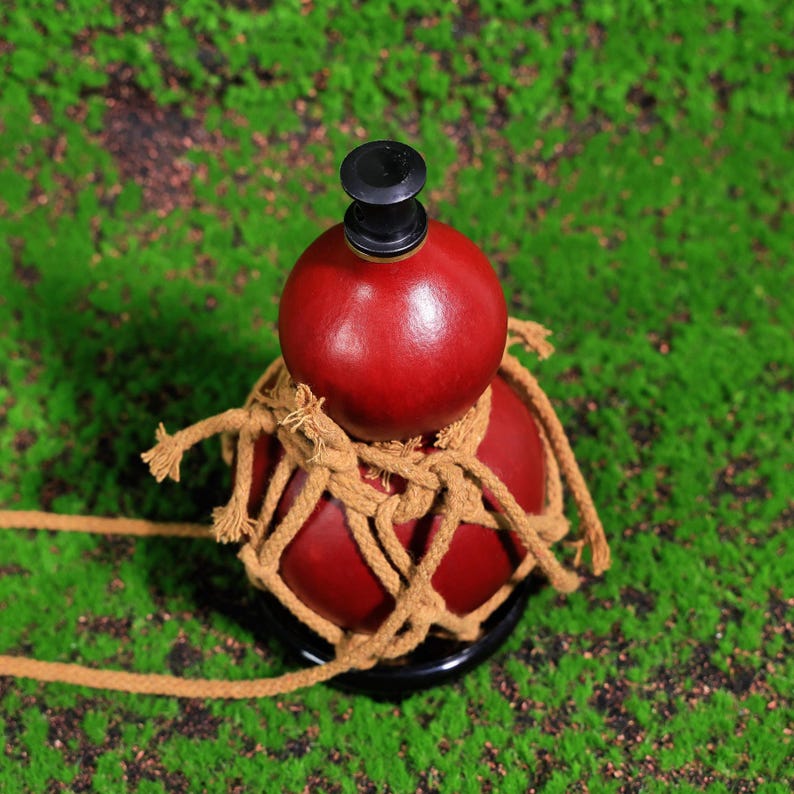 Puede incluir: Una botella de calabaza roja con un tap&oacute;n negro, envuelta en cuerda marr&oacute;n. La botella est&aacute; sobre una superficie verde.
