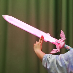 Puede incluir: Una espada de juguete de pl&aacute;stico rosa y azul con una hoja rosa brillante. La espada est&aacute; siendo sostenida por la mano de una persona.