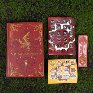 May include: A collection of Final Fantasy XIV merchandise, including a red book with gold accents and a dragon design, a bookmark, and a sheet of stickers featuring various character designs. The items are arranged on a green surface.