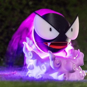 Może przedstawiać: Wydrukowana w 3D rzeźba postaci Pokémon Gastly. Rzeźba ma czarne ciało, białe oczy i czerwone usta z kłami. Emituje fioletową poświatę i jest otoczona podstawą w kształcie chmury, również w kolorze fioletowym.