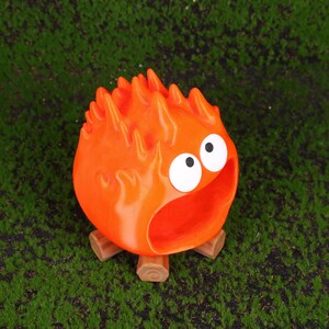 Fire Demon Calcifer-inspired desktop organizer made using high-quality 3D printing technology, placed on a clean modern workspace. The vibrant orange figure is styled to resemble Calcifer from Howl’s Moving Castle, with an open mouth serving as a functional storage compartment. Ideal for holding small desk accessories like USB drives, SD cards, or paper clips. This product blends whimsical anime charm with practical organization for anime lovers and Studio Ghibli fans.