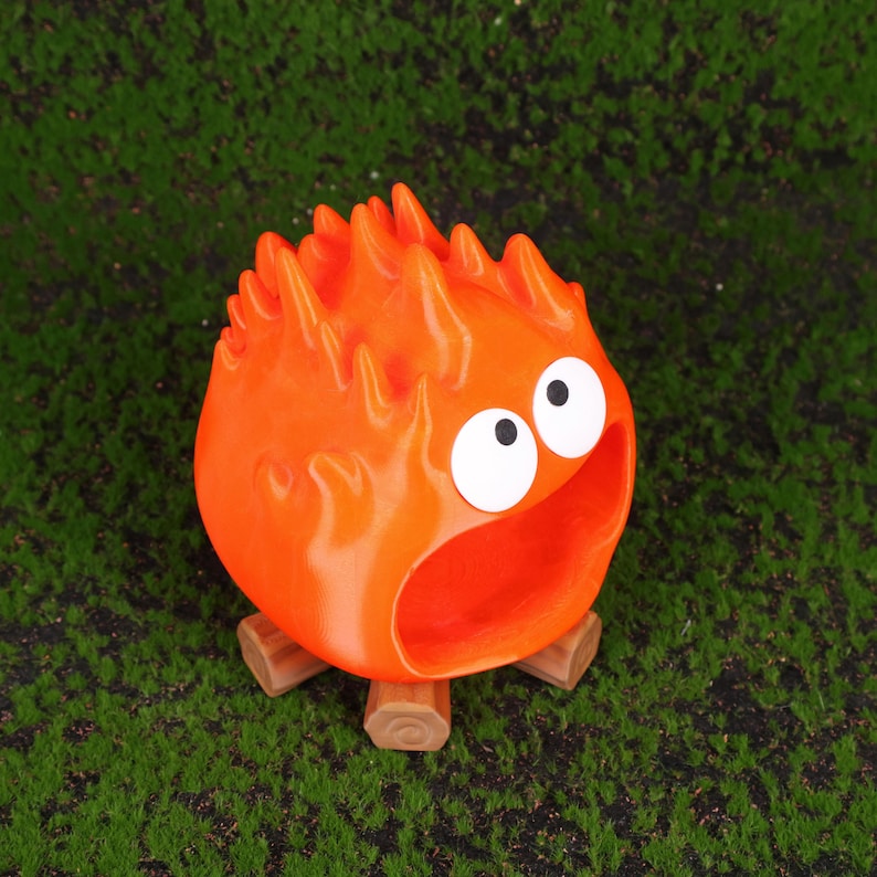 Fire Demon Calcifer-inspired desktop organizer made using high-quality 3D printing technology, placed on a clean modern workspace. The vibrant orange figure is styled to resemble Calcifer from Howl’s Moving Castle, with an open mouth serving as a functional storage compartment. Ideal for holding small desk accessories like USB drives, SD cards, or paper clips. This product blends whimsical anime charm with practical organization for anime lovers and Studio Ghibli fans.