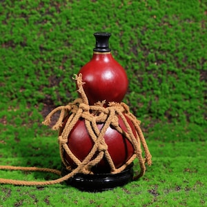 Puede incluir: Una botella de calabaza roja con un tap&oacute;n negro, envuelta en una red de cuerda marr&oacute;n, sentada sobre una base de madera sobre un fondo de musgo verde.