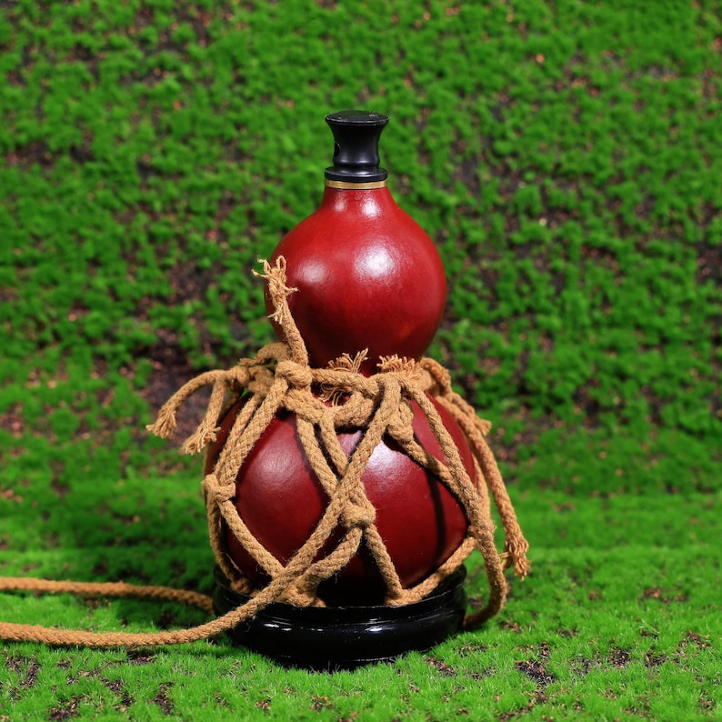 Puede incluir: Una botella de calabaza roja con un tap&oacute;n negro, envuelta en una red de cuerda marr&oacute;n, sentada sobre una base de madera sobre un fondo de musgo verde.