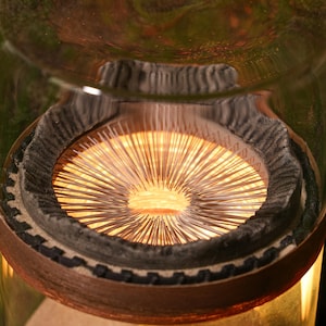 Può includere: Primo piano di un apparecchio di illuminazione circolare con un anello di piccole aste metalliche sottili che irradiano verso l'esterno dal centro. L'apparecchio di illuminazione è racchiuso in un cilindro di vetro trasparente e circondato da un anello di legno marrone scuro.