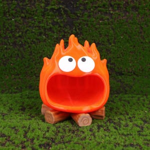 Studio Ghibli-themed Calcifer fire demon organizer for desk storage displayed on a wooden tabletop next to a laptop and stationery. The design captures Calcifer’s playful expression with bright colors and smooth 3D printed detailing. Perfect for organizing small items at home or the office, this Howl’s Moving Castle-inspired decor adds a magical touch to your workspace while serving as a thoughtful gift for Miyazaki fans.
