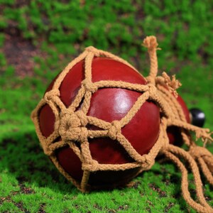 Puede incluir: Una calabaza roja con una red de cuerda tejida, la calabaza est&aacute; atada con una cuerda gruesa y tiene un tap&oacute;n de madera marr&oacute;n.