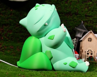 May include: A green and white 3D printed figurine of a Pokemon character, Bulbasaur, with its mouth open and eyes closed. The figurine is sitting on a green surface.