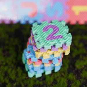 May include: A stack of interlocking foam puzzle floor tiles with a textured surface. The top tile is light green with a purple number two printed in the center. The other tiles in the stack are various pastel colors including yellow, blue, and pink.