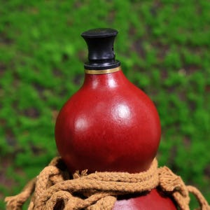 Puede incluir: Una botella de calabaza roja con un tap&oacute;n negro y dorado. La botella est&aacute; atada con una cuerda marr&oacute;n.