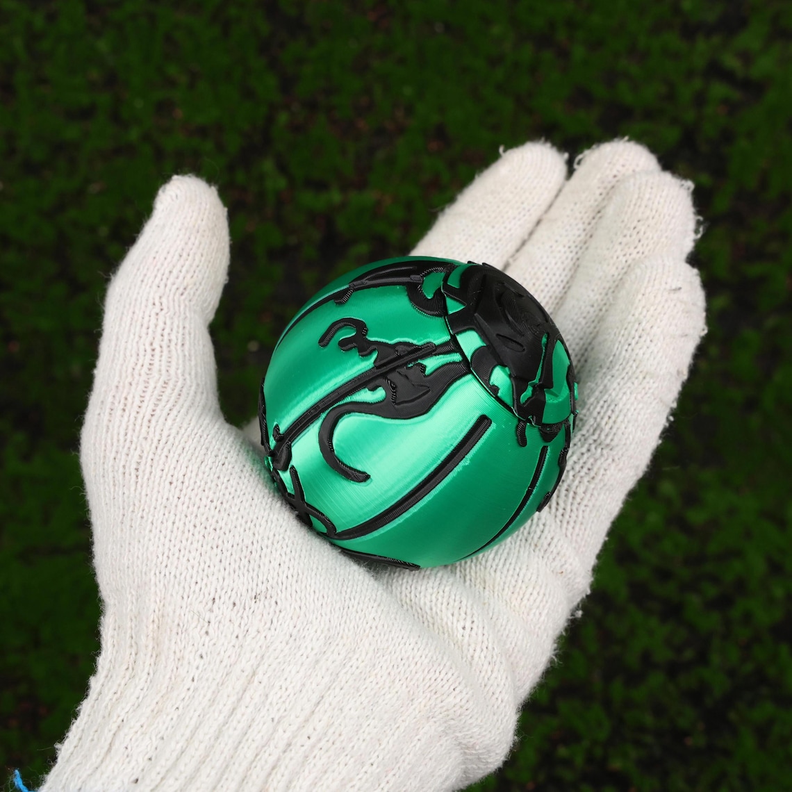 May include: A green and black sphere with a detailed, patterned design. The sphere is held in a white gloved hand against a blurred green background. The sphere's design features curved lines and shapes.
