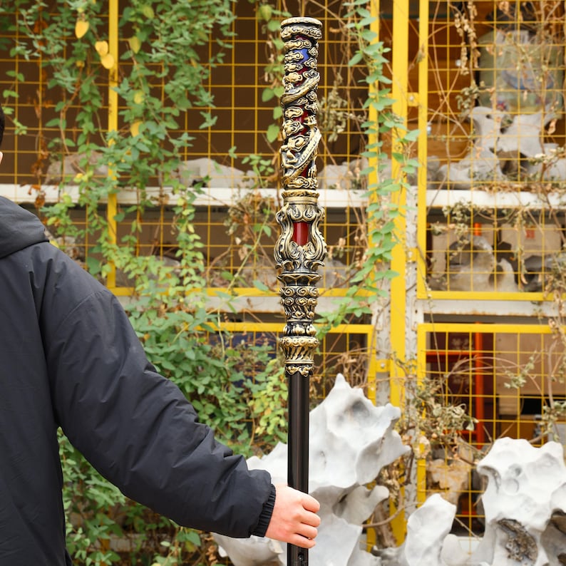 May include: Ornate staff with intricate carvings and a dark shaft. The top section features a detailed design with gold, red, and blue accents. The staff appears to be made of metal and is held by a person.