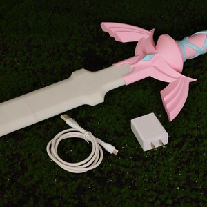 Puede incluir: Una espada de pl&aacute;stico rosa y blanca con un cable de carga blanco y un adaptador de pared blanco.