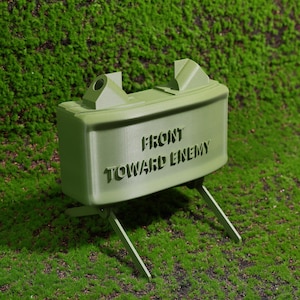 May include: A green plastic model of a military anti-personnel mine with the words "FRONT TOWARD ENEMY" printed on the side.