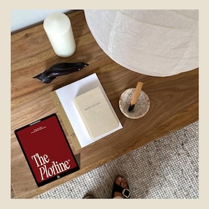May include: A wooden table with a white candle, a small wooden sculpture, a white paper lantern, a small book titled "Meditation", a small bowl with a stick of incense, and a tablet with the text "The Plotline" on the screen.