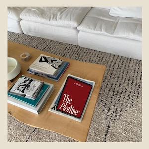 May include: A wooden coffee table with a stack of books and a magazine titled "The Plotline" on top. The table is on a light grey rug with a black and white pattern.
