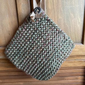 May include: A knitted, square pot holder in green and red hues, suspended from a wooden door. The textured surface and loop and button fastening give it a rustic, handcrafted feel, perfect for a country kitchen.