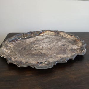 May include: An oval silver-toned serving tray featuring a scalloped, wavy edge and a tarnished, textured surface with visible dark oxidation patterns across the center and along the decorative, raised rim of the metal platter.
