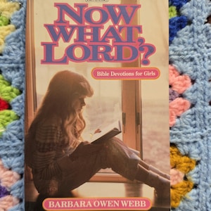 May include: A paperback book titled Now What, Lord? Bible Devotions for Girls by Barbara Owen Webb. The cover features an illustration of a person with long hair sitting by a window reading a book, with purple and pink text on a cream background.