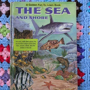 May include: A hardcover book titled A Golden Fun-To-Learn Book THE SEA AND SHORE. The cover features a large shark, various fish, a starfish, and underwater scenery. A green text box lists tide pools, shells, and other ocean wonders.