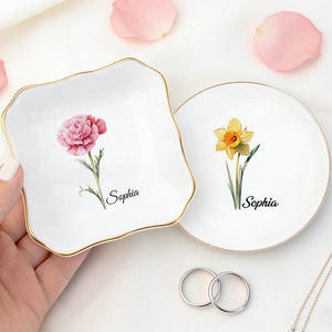 May include: Two white ceramic trinket dishes, one square and one round, with gold rims. Each dish features a floral design and the name "Sophia". Pink flower petals are scattered around the dishes. A silver necklace and rings are also visible.