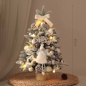 Könnte beinhalten: Ein kleiner, künstlicher Weihnachtsbaum, etwa 45 cm hoch, geschmückt mit Ornamenten, Lichtern und einer Schleife. Der Baum hat Tannenzapfen, silberne und klare Kugeln und ein "Merry Christmas"-Schild. Eine kleine Engelsfigur ist ebenfalls enthalten.