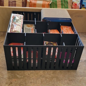 May include: A black plastic organizer with 12 compartments, filled with various small packets of condiments. The organizer has a grid pattern and is sitting on a brown surface. The text "ENJOY RESPONSIBLY" is visible on a blue packet.