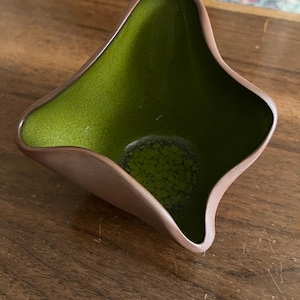 May include: A small, square-shaped ceramic bowl with a unique design. The interior is a vibrant green, while the exterior rim is a warm brown. The bowl's edges curve gently, creating a distinctive, eye-catching shape. The bowl is empty.
