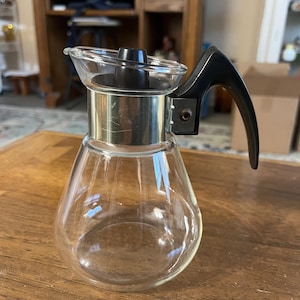 May include: A clear glass carafe with a wide, rounded base and a tapered neck. It features a metallic silver band around the upper section, a black plastic handle attached with a visible screw, and a black plastic lid insert at the top opening.