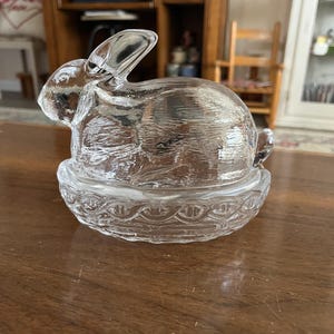 May include: A clear glass bunny figurine sitting atop a decorative glass dish. The bunny is detailed with textured fur and upright ears. The dish has a scalloped edge design. The figurine is on a wooden surface.