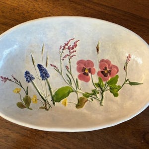 May include: Oval-shaped decorative bowl with a light-colored background. The bowl is adorned with a floral design featuring pink pansies, blue grape hyacinths, and other wildflowers. The flowers are painted in detail, adding a touch of nature.