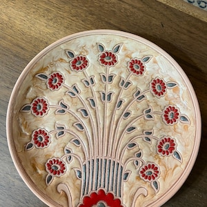 May include: A decorative ceramic plate with a floral design. The plate features a beige background with a pattern of red flowers and gray leaves. The rim of the plate is a light pink color.