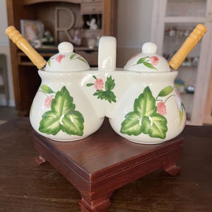 May include: A white ceramic double jar set with green leaves and pink berries. The jars have lids and come with two wooden spoons. The set is on a wooden base.