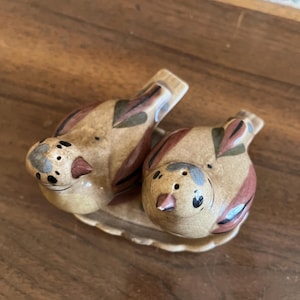 May include: A ceramic salt and pepper set featuring two bird-shaped shakers resting on a matching dish. The birds are painted in shades of tan, brown, and red, with black and blue accents. The set is designed for tabletop use.