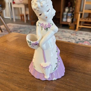 May include: A vintage ceramic figurine of a woman in a white dress with a lavender apron and skirt. The figurine is holding a basket and a small container, both with lavender accents. The woman has her eyes closed and is wearing a floral design.