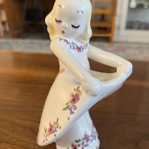 May include: A vintage ceramic figurine of a girl with closed eyes and blonde hair, wearing a white dress adorned with pink and blue floral accents. The figurine is holding a white, curved object, possibly a basket or a decorative element.
