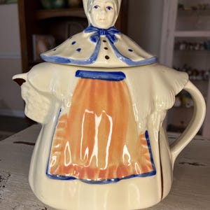 Puede incluir: Tetera de cerámica color crema con tapa que representa el rostro de una mujer y un gorro floral. La tetera tiene un asa y está decorada con detalles azules y naranjas, incluido un diseño de delantal azul. Un artículo de cocina vintage.