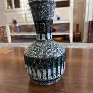 May include: A ceramic vase with a speckled black and light blue finish. The body features vertical ridges with alternating dark green and white panels. The neck is narrow and cylindrical, transitioning into a wider, rounded base with a textured surface.