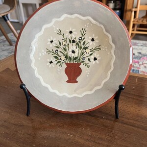 May include: A round, hand-painted ceramic plate with a floral design. The plate features a red rim, a gray border, and a cream-colored center with a vase of white daisies and black accents. It is displayed on a black metal stand.