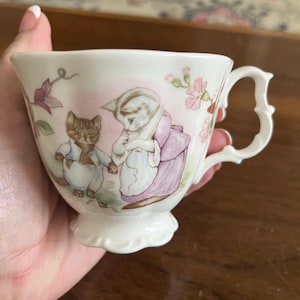 May include: White teacup with a decorative handle and a scalloped base. The cup features a charming illustration of two cats in clothing, with pink and purple floral accents. The cup is held in a hand.