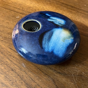 May include: A round, blue ceramic flower frog with a glossy finish. The frog has a circular opening with a metal pin holder for arranging flowers. The glaze features shades of blue and white, creating a marbled effect.