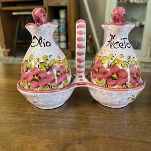 May include: A ceramic oil and vinegar set with a pink and white speckled base. The bottles are decorated with red flowers and the words "olio" and "aceto". The set includes a handle with red accents.