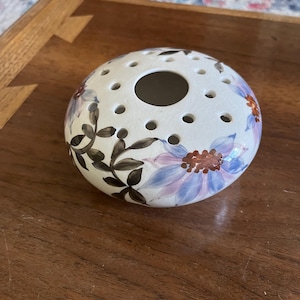 May include: A white ceramic flower frog with a round shape and multiple holes. The frog is decorated with blue and purple floral designs and brown leaves. The center has a large opening for holding flowers. The frog sits on a wooden surface.