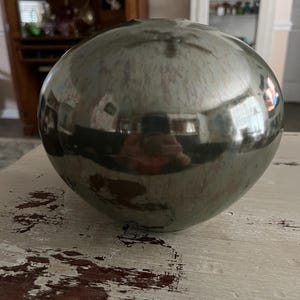 May include: A spherical, glazed ceramic vase with a reflective, metallic finish. The vase is a muted green-gray color and has a smooth, rounded shape. The surface reflects the surrounding environment, creating a mirrored effect. The vase is a decorative home accent.