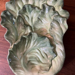 May include: A ceramic serving dish shaped like a head of lettuce. The dish is painted in shades of green and white, with detailed leaf textures. The dish is designed to resemble a realistic head of lettuce.