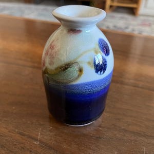 May include: A small, decorative ceramic vase with a white rim and a rounded body. The vase features a color gradient from dark blue to white, with floral accents in shades of pink, green, and blue. The vase is sitting on a wooden surface.
