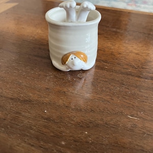 May include: A small, off-white ceramic container with a whimsical design. The container features a face with orange hair and two white mushroom-shaped objects inside. The container is sitting on a brown wooden surface.