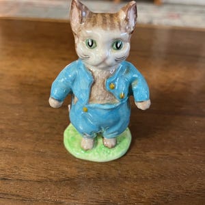 May include: A ceramic figurine of a cat wearing a blue outfit with gold buttons, standing on a green base. The cat has brown and tan fur with green eyes. The figurine is a decorative item.
