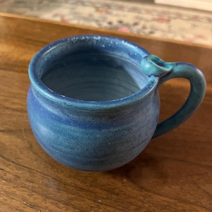 May include: A handmade, blue ceramic mug with a rounded body and a curved handle. The mug has a textured surface and a gradient of blue hues, from a lighter shade at the top to a deeper tone at the base. The mug is sitting on a wooden surface.